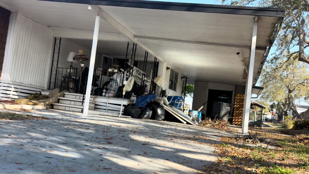 A cluttered porch with scattered old furniture, bags of trash, and debris, under a shaded roof in a residential area.