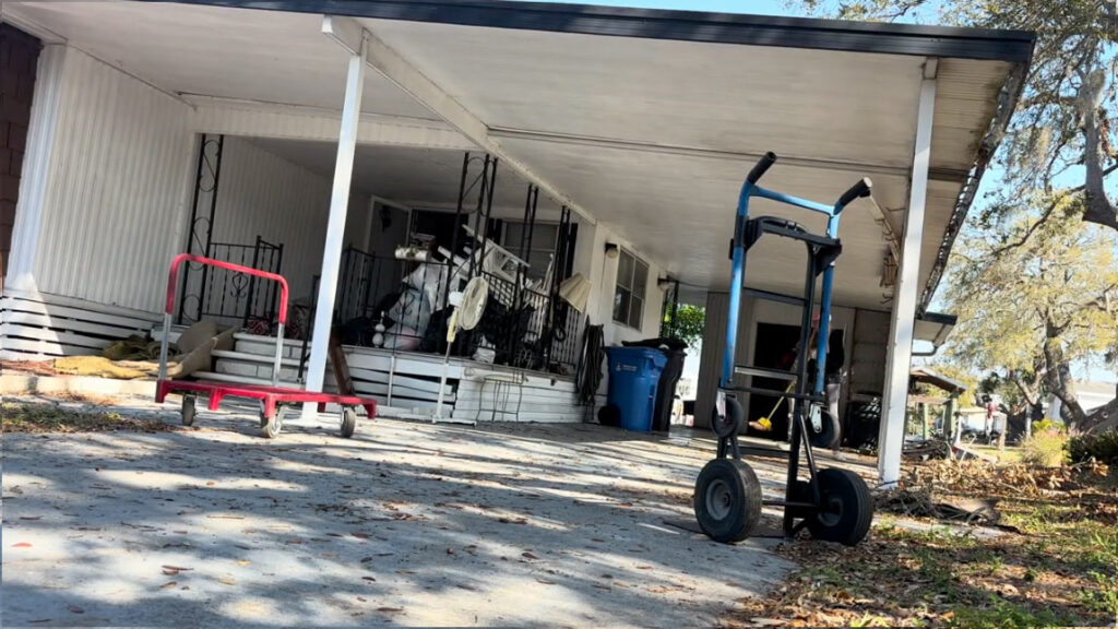 Apollo Beach House Cleanout 3 A blue hand truck stands on a driveway, with a red cart nearby and a cluttered porch in the background under a covered area.