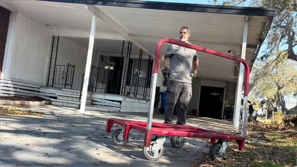 Apollo Beach House Cleanout 2 A person stands on a red utility cart outside a house, surrounded by a driveway and lawn with leaves scattered.