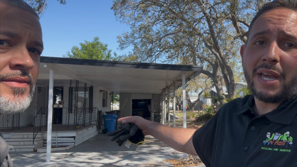 Apollo Beach House Cleanout 1 A person gestures towards a house with a covered porch, surrounded by trees, with a blue trash bin nearby.