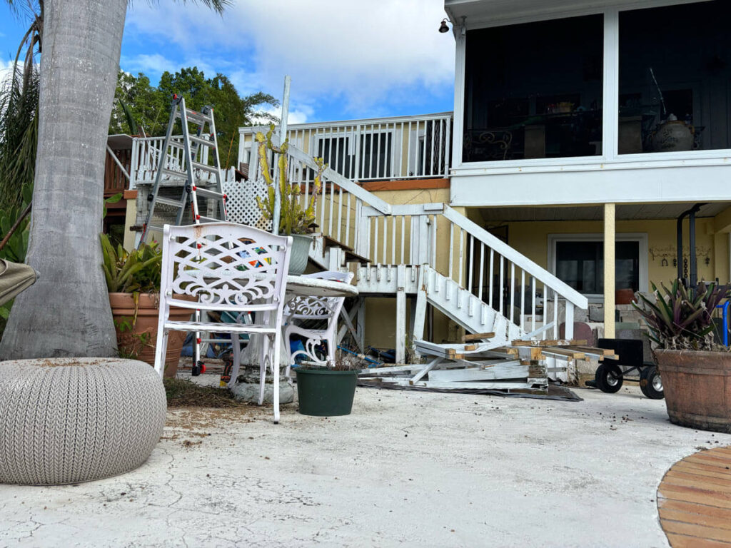 Bradenton Outdoor Staircase Demolition 3 Cluttered outdoor area with construction materials, a ladder, white chairs, and potted plants leading to a screened porch.
