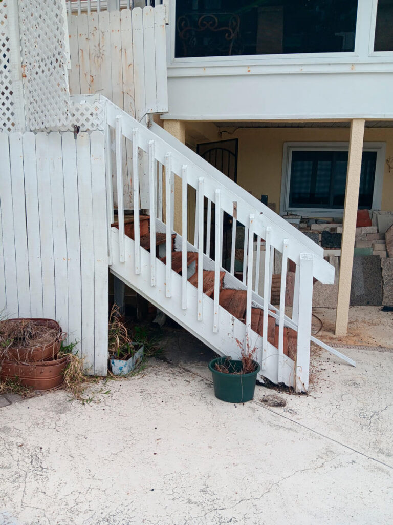 Bradenton Outdoor Staircase Demolition 2 A weathered white staircase leads up from a concrete patio, flanked by scattered potted plants and a wooden fence.
