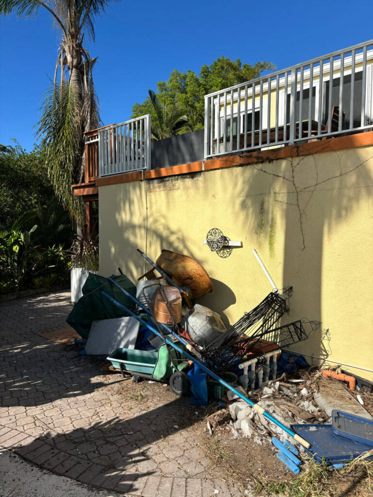 Bradenton Outdoor Staircase Demolition 9 A messy area with discarded items, including a wheelbarrow, pots, and debris, against a yellow wall and blue sky.