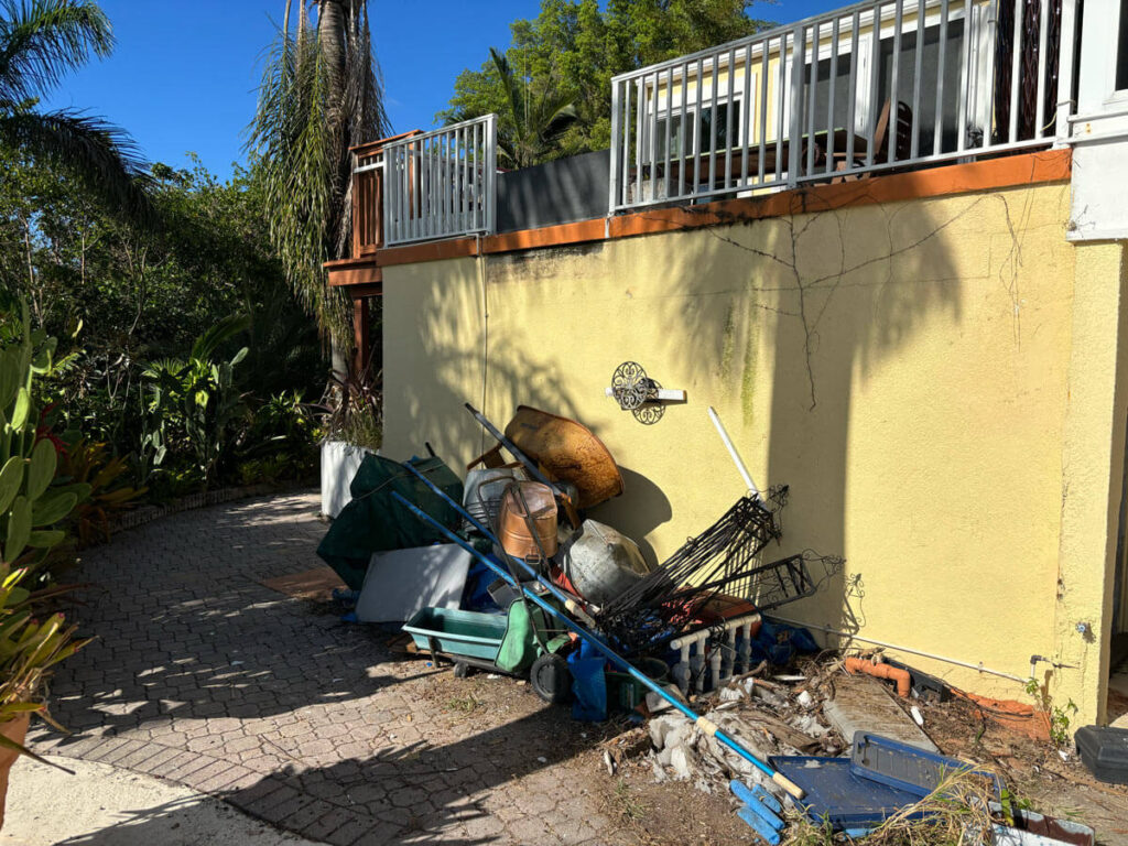 Bradenton Outdoor Staircase Demolition 7 A pile of discarded items, including buckets and old furniture, is amassed against a yellow wall, surrounded by lush greenery.