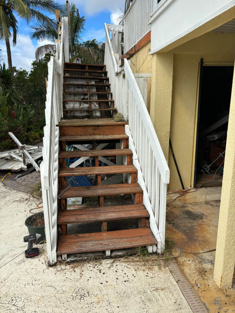 Bradenton Outdoor Staircase Demolition 6 Weathered wooden stairs lead up to a raised structure, surrounded by tropical plants and debris, under a partly cloudy sky.