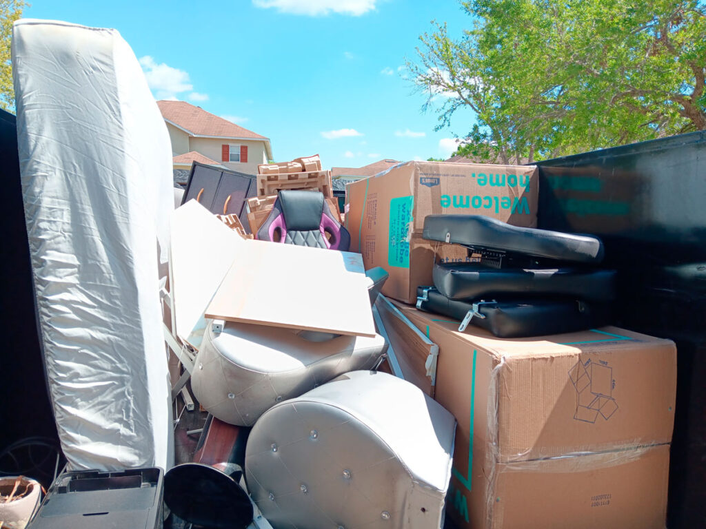 A cluttered dumpster filled with assorted furniture, boxes, and office chairs under a bright blue sky.