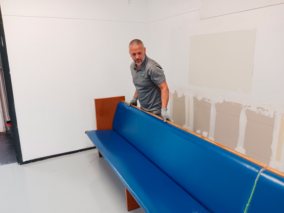 A person lifts a blue padded bench in a sparsely decorated room with unfinished walls and a polished floor.