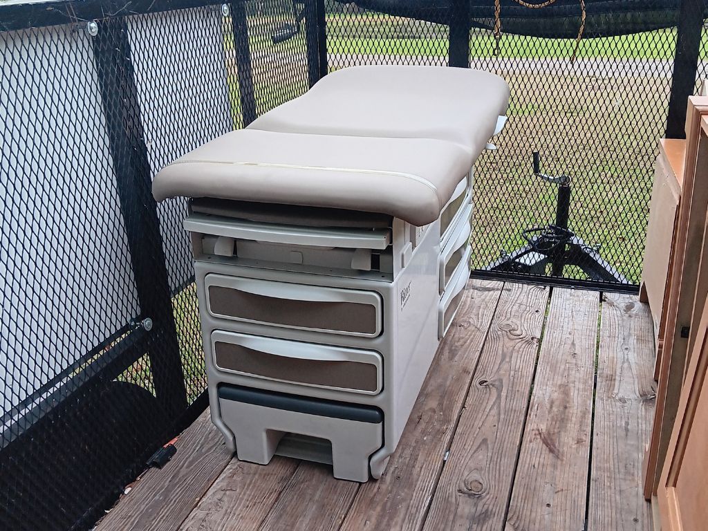 A portable medical exam table with a beige cushion sits on a wooden deck, surrounded by a black safety railing and green landscape.