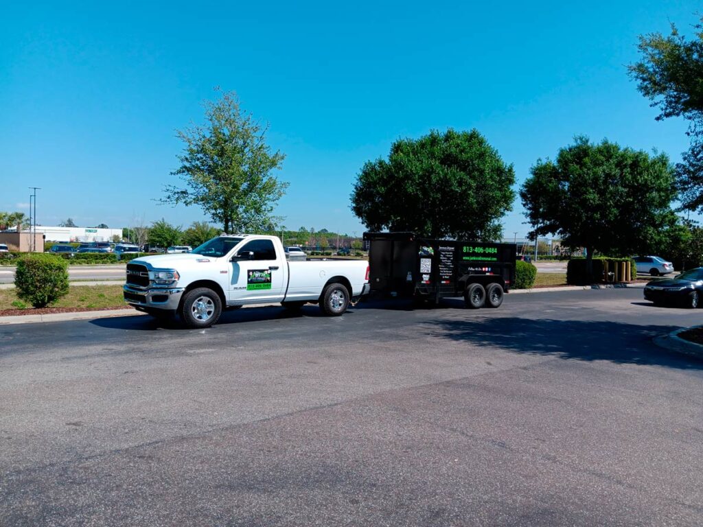 House Cleanout in Riverview 3 A white pickup truck towing a black trailer parked on a sunny day, flanked by green trees and a clear blue sky.