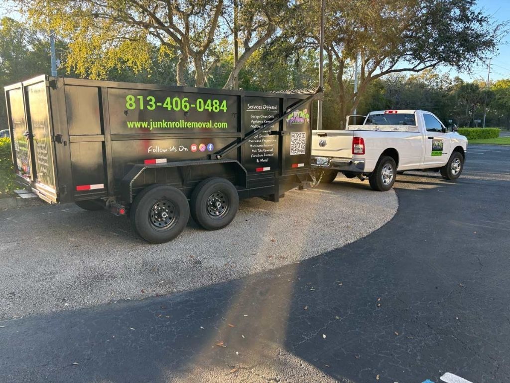 Big News: Junk N Roll Just Got Even Bigger & Better! 2 A black junk removal trailer hitched to a white pickup truck, both parked in a tree-lined area with clear signage promoting the service.
