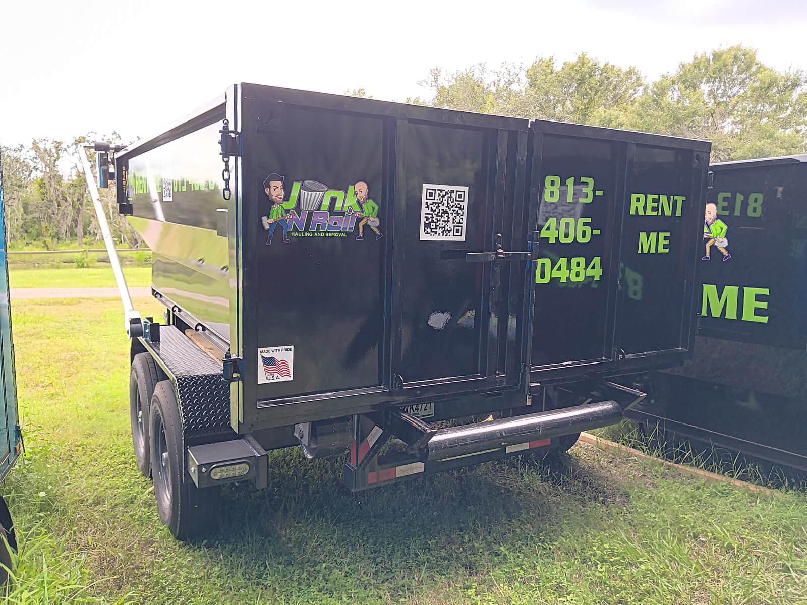 An angled view of a black Junk N Roll hauling and removal dumpster, showing '813-406-0484', 'RENT ME', the company logo, and a QR code, set against a backdrop of trees and sky.