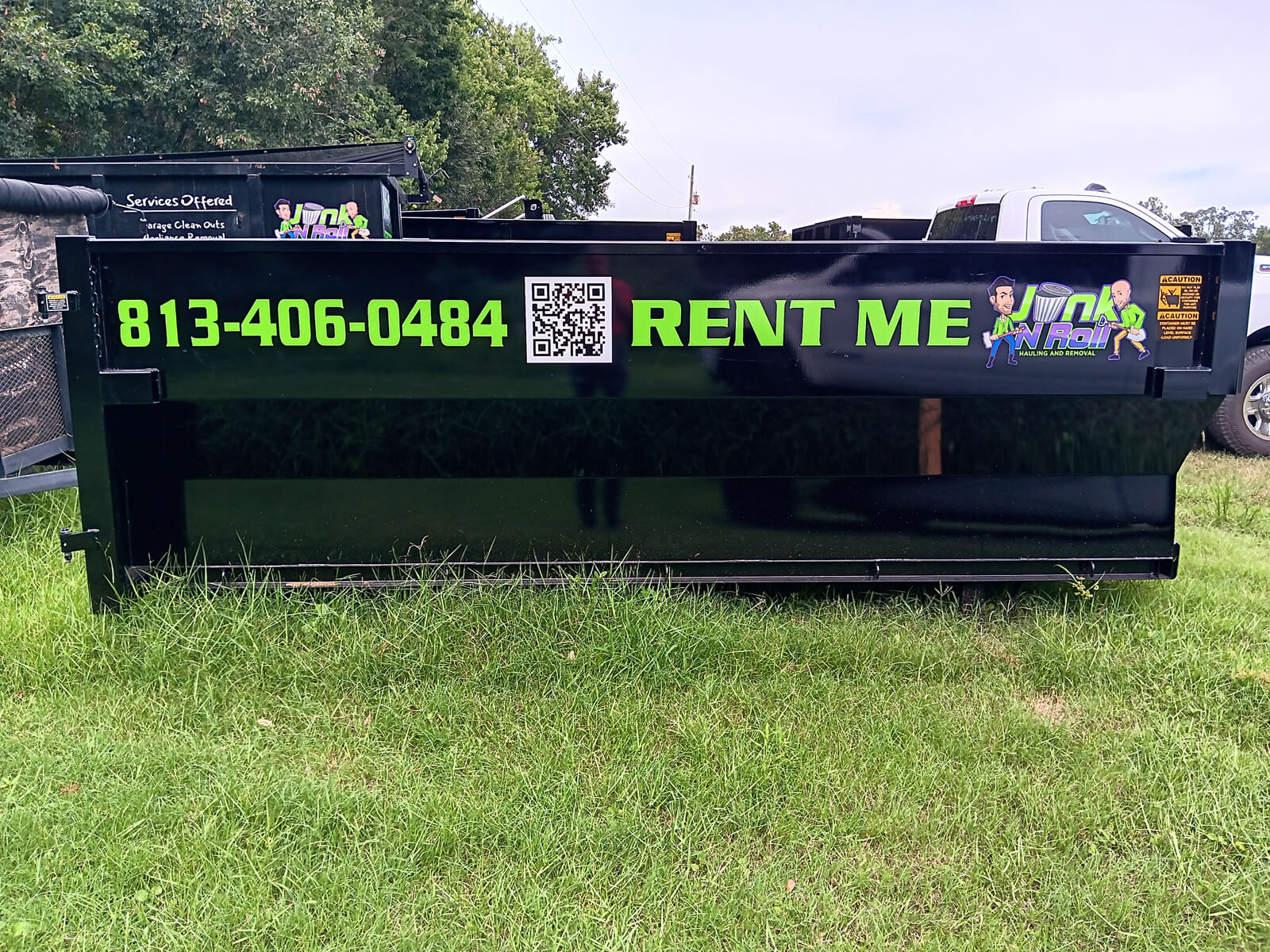 A black Junk N Roll hauling and removal dumpster with 'RENT ME' and the phone number '813-406-0484' in green lettering, along with the company logo and a QR code, parked on grass with trees in the background.