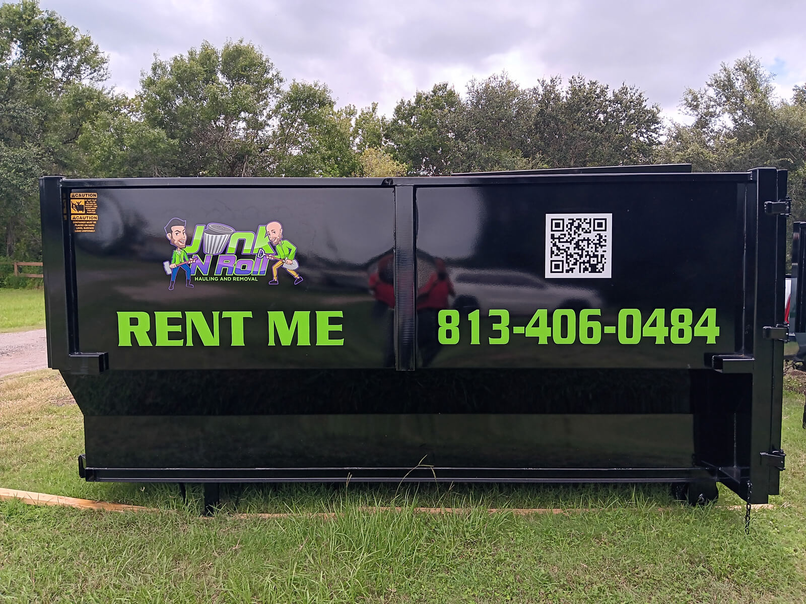 A black Junk N Roll hauling and removal dumpster with 'RENT ME' and the phone number '813-406-0484' in green lettering, the company logo, and a QR code, set against a green treeline.