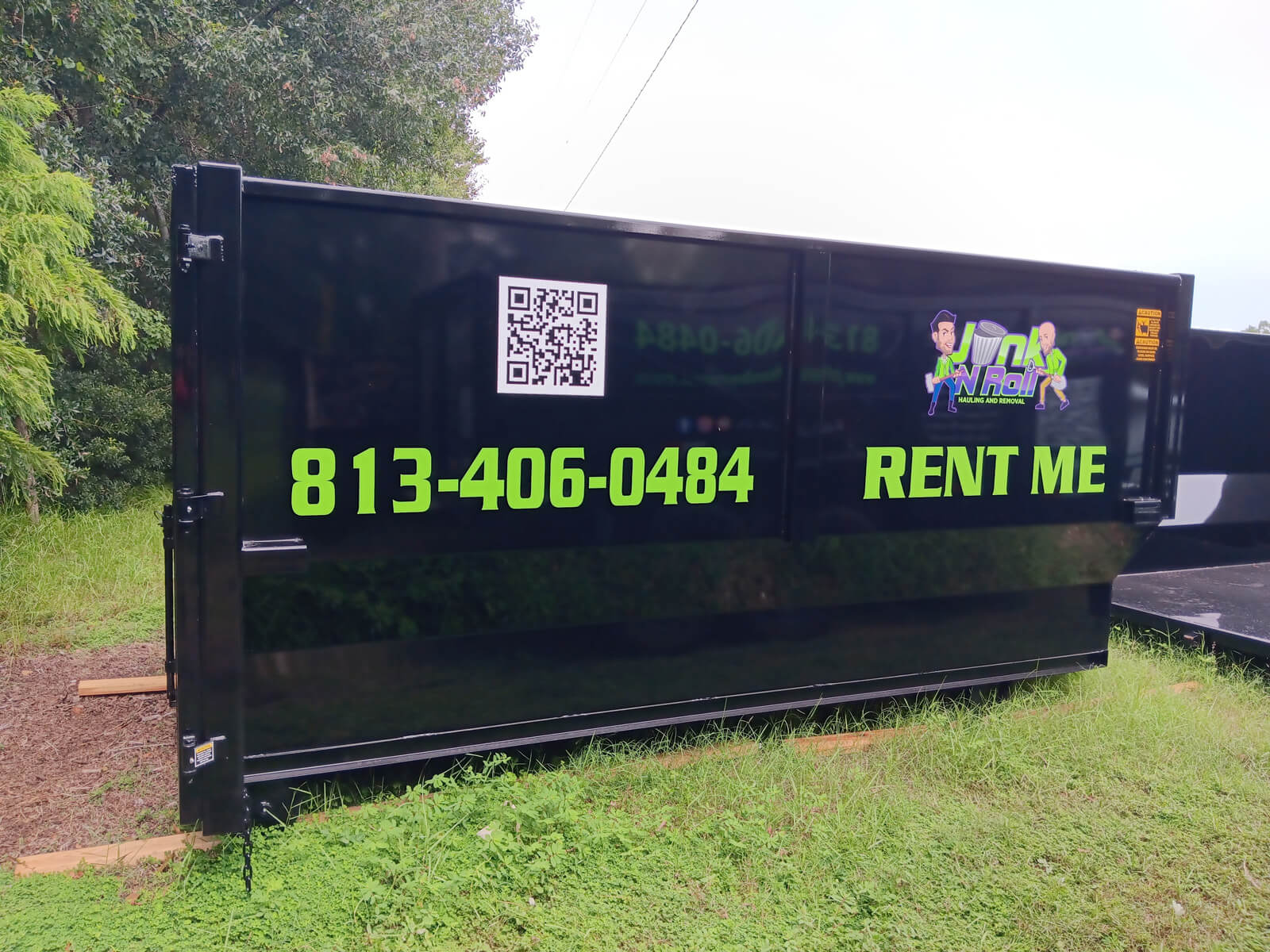 A black Junk N Roll hauling and removal dumpster seen from the side, displaying the phone number '813-406-0484', a QR code, 'RENT ME', and the company logo, with another dumpster partially visible in the background and a white truck.