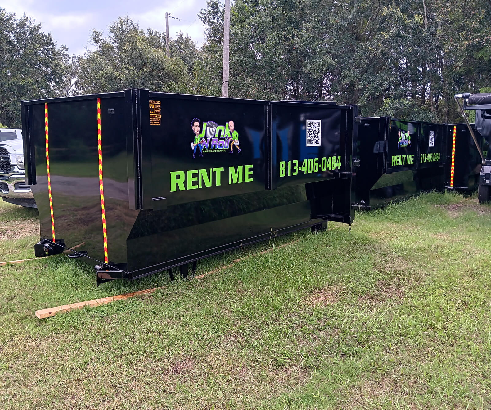 Two black Junk N Roll hauling and removal dumpsters side-by-side on a grassy field with trees in the background. The front dumpster displays 'RENT ME', the phone number '813-406-0484', the company logo, and a QR code, along with yellow and red reflective tape on its corners.