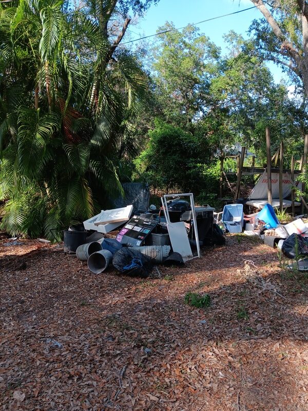 A large pile of junk and debris on a ground covered in dry leaves, with palm trees and other vegetation in the background.