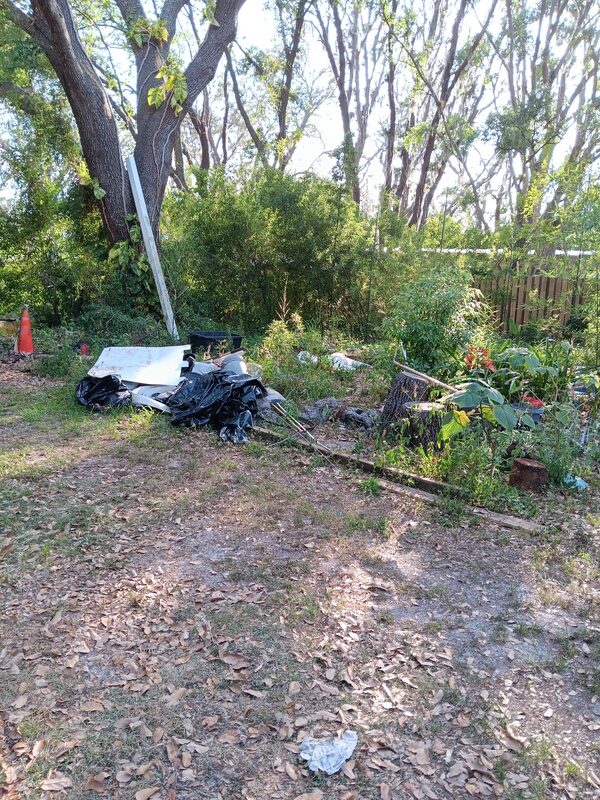 An overgrown yard with a large pile of black trash bags and a white sheet on the ground.