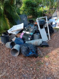 A large pile of junk and household trash, including a grill, cardboard boxes, and various plastic buckets, in a wooded yard.