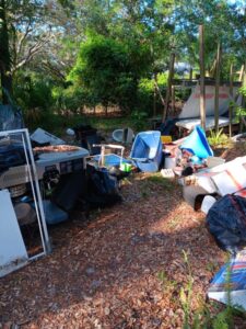 A pathway through a wooded area with trees overhead, surrounded by various discarded items and junk.