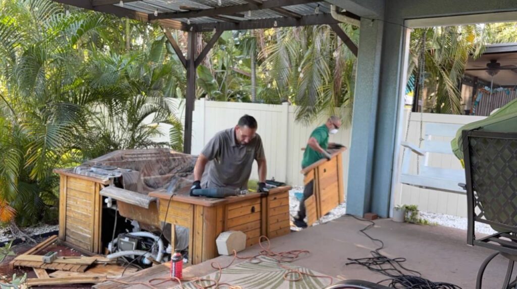 Two workers are carrying away a large wooden side panel from a disassembled hot tub.