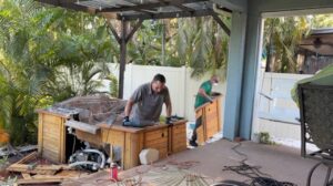 Two workers are carrying away a large wooden side panel from a disassembled hot tub.