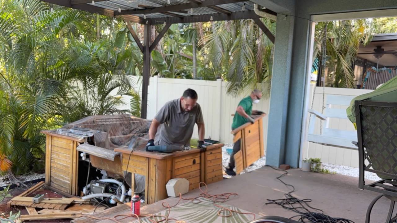 Home 10 Two workers are carrying away a large wooden side panel from a disassembled hot tub.