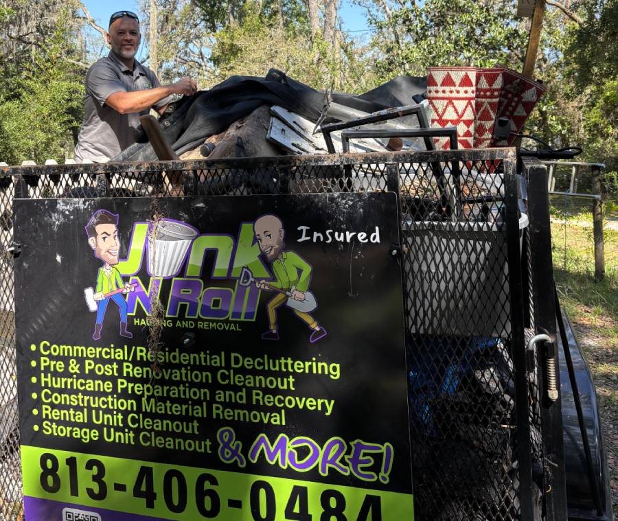 A man in a gray shirt standing behind a large trailer filled with junk. The side of the trailer has the logo for a junk removal company called "Junk N Roll."