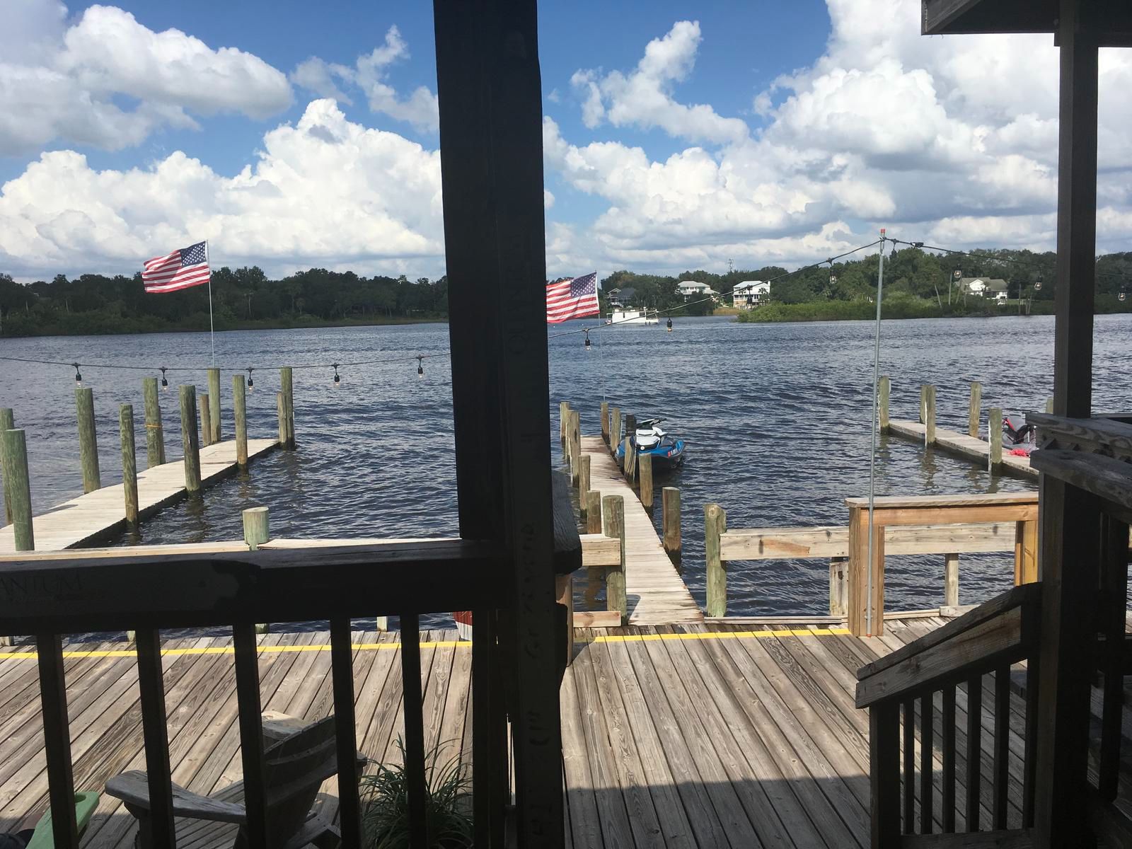 View of a peaceful waterway with docked boats, American flags waving, and fluffy clouds in the blue sky.