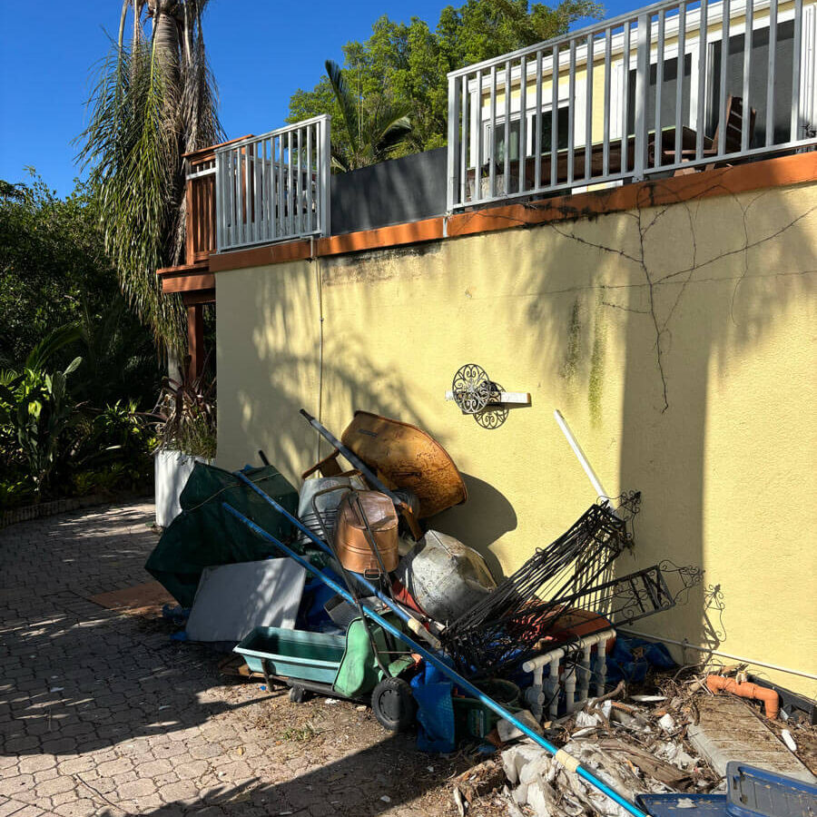 A messy area with discarded items, including a wheelbarrow, pots, and debris, against a yellow wall and blue sky.